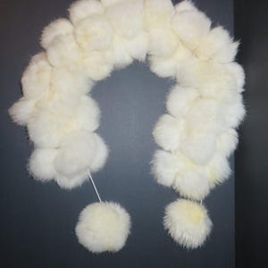May include: A fluffy, white fur scarf with a rounded shape. The scarf is made of soft, textured fur and features two small, round pom-poms attached at the ends. The scarf is displayed against a dark gray background.