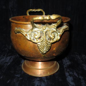 May include: A copper and brass planter with ornate handles. The planter has a round body and a flared base.