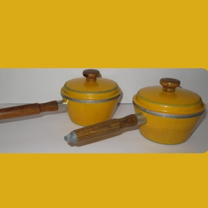 Puede incluir: Juego de utensilios de cocina de esmalte amarillo con asas y tapas de madera. El juego incluye una cacerola y dos ollas. Las asas y los pomos de las tapas son de madera. La vajilla tiene un ribete plateado.