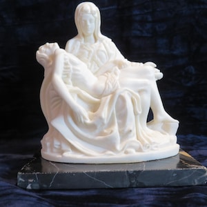 May include: A white marble sculpture depicting a seated figure holding a reclining figure in their lap, featuring intricate folds in the draped clothing. The sculpture is mounted on a rectangular base made of polished black marble with grey veining.