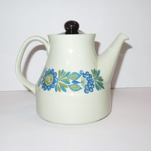 May include: A cream-colored ceramic teapot featuring a dark brown lid knob and a horizontal band of blue and green floral and leaf patterns encircling the body. The teapot has a curved handle and a tapered spout, with a smooth, glossy finish.
