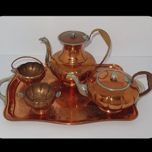 May include: A vintage copper tea set comprising a tray, teapot, creamer, and sugar bowl. The teapot features a curved handle and a lid with a silver knob. The set is presented on a copper tray with silver handles.