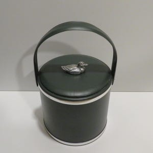 Pode incluir: Um balde de gelo verde escuro com uma estatueta de pato prateado na tampa e uma alça a condizer. O balde tem faixas prateadas na parte superior e inferior. A alça é arqueada e feita do mesmo material verde escuro.