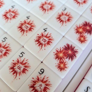 May include: A collection of white rectangular mahjong tiles featuring red firework illustrations. Each tile displays a black Arabic numeral in the corner and a corresponding red Chinese character centered within the firework design on the face.