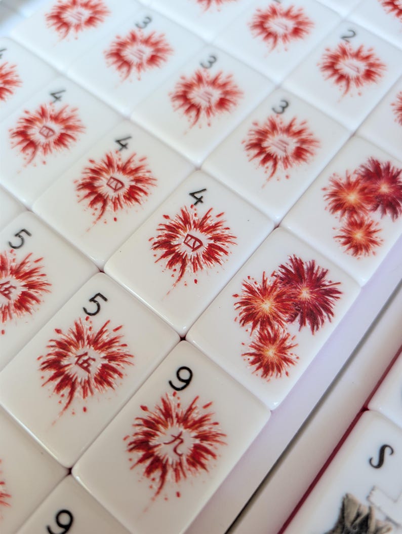 May include: A collection of white rectangular mahjong tiles featuring red firework illustrations. Each tile displays a black Arabic numeral in the corner and a corresponding red Chinese character centered within the firework design on the face.