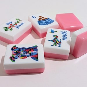 May include: A collection of white and pink Mahjong tiles. Some tiles feature colorful illustrations of a dog, flowers, and a bowl of food. Other tiles are solid pink. The word "north" is written on one tile.