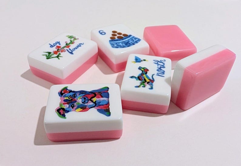 May include: A collection of white and pink Mahjong tiles. Some tiles feature colorful illustrations of a dog, flowers, and a bowl of food. Other tiles are solid pink. The word "north" is written on one tile.