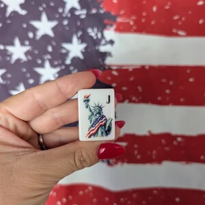 May include: A rectangular white mahjong tile featuring a colorful illustration of the Statue of Liberty draped in an American flag, with the letter J printed in black in the upper right corner. The tile has a smooth, glossy finish.