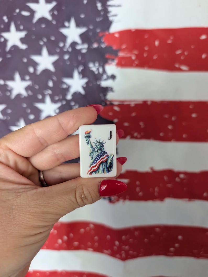 May include: A rectangular white mahjong tile featuring a colorful illustration of the Statue of Liberty draped in an American flag, with the letter J printed in black in the upper right corner. The tile has a smooth, glossy finish.