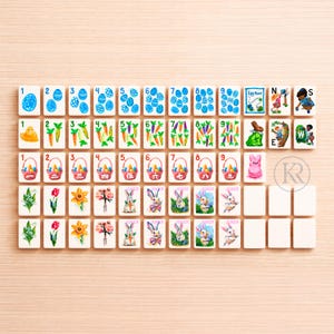 May include: A collection of Easter-themed Mahjong tiles. The tiles feature images of Easter eggs, carrots, baskets, flowers, and bunnies. The tiles are arranged on a light wood surface.
