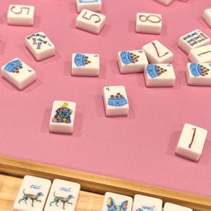 May include: A collection of white Mahjong tiles with various illustrations, including numbers, dog bowls, and dog portraits. The tiles are scattered on a pink surface, with some arranged in a wooden tray.