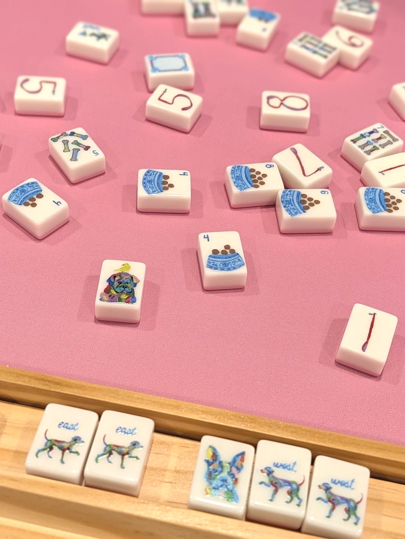 May include: A collection of white Mahjong tiles with various illustrations, including numbers, dog bowls, and dog portraits. The tiles are scattered on a pink surface, with some arranged in a wooden tray.