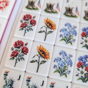 May include: A collection of rectangular white mahjong tiles featuring various printed designs including red roses, yellow sunflowers, blue forget-me-nots, the Statue of Liberty with a flag, and military boots with an American flag and the letter S.