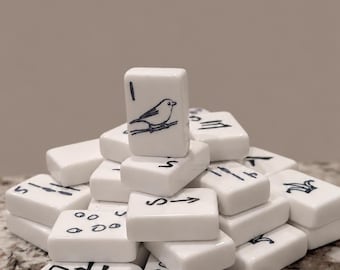 May include: A stack of white rectangular mahjong tiles with rounded corners. The tiles feature black ink illustrations of birds, numbers, and various symbols. One tile is positioned vertically at the top, displaying a bird perched on a branch.