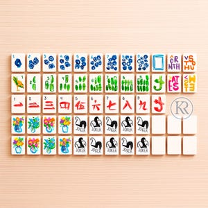 May include: A set of white Mahjong tiles with hand-painted designs. The tiles feature various symbols, numbers, and characters in blue, green, red, and black. Some tiles display floral arrangements, while others have the word "Joker".
