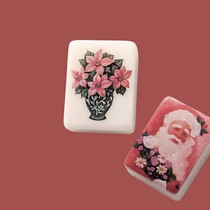 May include: Two rectangular decorative soaps. One features a pink poinsettia floral arrangement in a black vase. The other soap has a Santa Claus design with a pink and white colour scheme. Both soaps are on a red background.