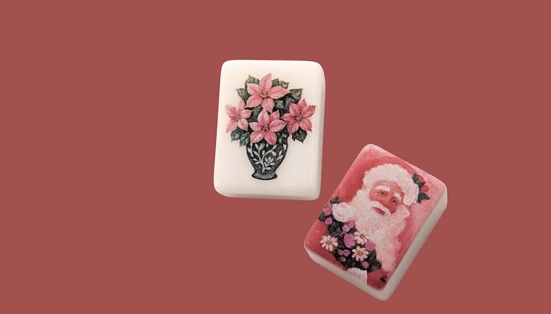 May include: Two rectangular decorative soaps. One features a pink poinsettia floral arrangement in a black vase. The other soap has a Santa Claus design with a pink and white colour scheme. Both soaps are on a red background.