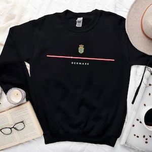 May include: Black sweatshirt with a red and white stripe across the chest and a small embroidered image of the Danish flag with the word "DENMARK" underneath.