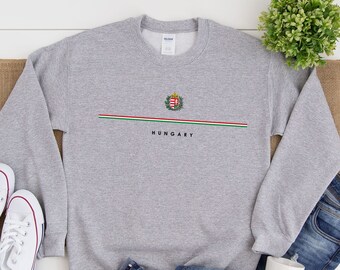 Puede incluir: Una sudadera gris con una franja blanca y verde a través del pecho. La franja presenta el escudo de armas húngaro y la palabra "HUNGARY" en texto negro.