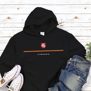 May include: Black hoodie with a white stripe and the word "Lithuania" printed on it. The hoodie has a yellow and red stripe above the word. A small coat of arms is above the stripe. The hoodie is laid out on a white wooden surface with a pair of white sneakers and blue jeans.