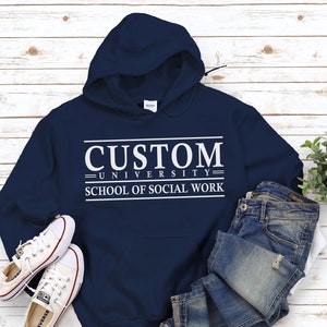 Może przedstawiać: Bordowa bluza z kapturem z białym nadrukiem, na którym widnieje napis "CUSTOM UNIVERSITY SCHOOL OF SOCIAL WORK". Bluza leży na białym drewnianym tle, obok niej znajdują się białe trampki i niebieskie dżinsy.
