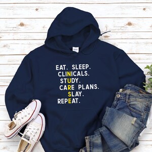 Può includere: Una felpa con cappuccio blu navy con una grafica gialla che recita "EAT. SLEEP. CLINICALS. STUDY. CARE PLANS. SLAY. REPEAT."