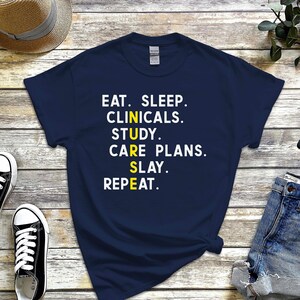 Può includere: T-shirt blu navy con testo bianco e giallo: "EAT. SLEEP. CLINICALS. STUDY. CARE PLANS. SLAY. REPEAT." La maglietta è abbinata a pantaloncini di jeans, scarpe da ginnastica bianche e nere, un cappello di paglia e uno smartwatch.