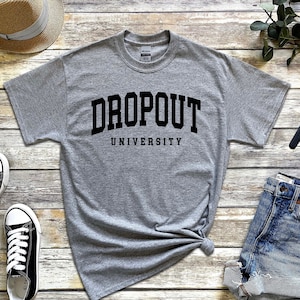 May include: Gray t-shirt with black text that reads "DROPOUT UNIVERSITY". The shirt is laid flat on a wooden surface with a pair of black sneakers, a straw hat, a pair of denim shorts, a watch, and earbuds.