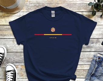 May include: Navy blue t-shirt featuring the Spanish coat of arms and the word "SPAIN" in yellow. A horizontal line in the colors of the Spanish flag is above the text. The shirt is displayed with jeans, sneakers, and a hat.