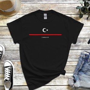 May include: Black t-shirt with a white crescent moon and star design and the word "TURKEY" printed below. The shirt is laid flat on a wooden surface with a straw hat, denim shorts, sneakers, and a watch.