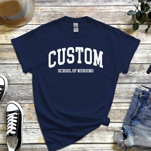 以下が含まれることがあります： 白い文字で「CUSTOM SCHOOL OF NURSING」と書かれた紺色のTシャツです。