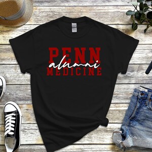 May include: Black t-shirt with red lettering that says "Penn alumni medicine" in a white script font.