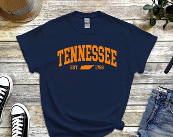 May include: Navy blue t-shirt with the word "TENNESSEE" in orange arched lettering. Below the word is "EST. 1796" with a Tennessee state outline. The shirt is displayed with a pair of black sneakers, a straw hat, and denim shorts.