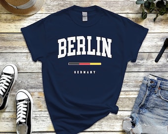 May include: A navy blue t-shirt with white text that reads "BERLIN" in a collegiate font. Below the text is a black, red, and yellow horizontal stripe representing the German flag, and the word "GERMANY" in white text.