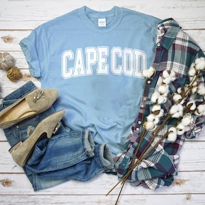 May include: A light blue t-shirt with the text "CAPE COD" in white block letters. The shirt is laid out on a white wooden surface with a pair of brown suede loafers, a pair of blue jeans, and a plaid shirt with a sprig of cotton.