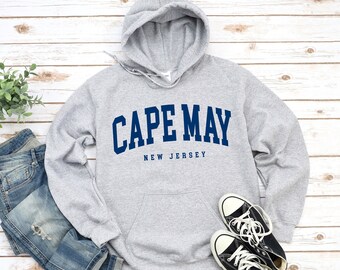 May include: A grey hooded sweatshirt displaying the text "CAPE MAY NEW JERSEY" in navy blue. The sweatshirt features a front pocket and a drawstring hood. Blue jeans and black trainers are also visible in the image.