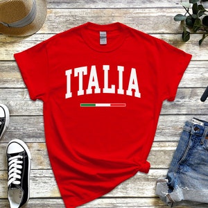 May include: Red t-shirt with white text that says "ITALIA" and a small green, white, and red stripe underneath. The shirt is laid out on a wooden surface with a straw hat, blue denim shorts, and black sneakers.