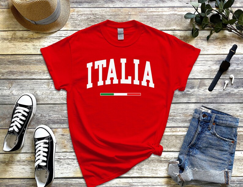 May include: Red t-shirt with white text that says "ITALIA" and a small green, white, and red stripe underneath. The shirt is laid out on a wooden surface with a straw hat, blue denim shorts, and black sneakers.