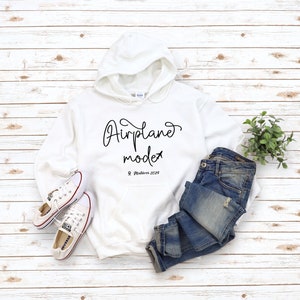 May include: White hoodie with the text "Airplane mode" and a small airplane icon. Below the text is "Maldives 2020". The hoodie is laid out on a white wood surface with a pair of white sneakers and blue jeans.