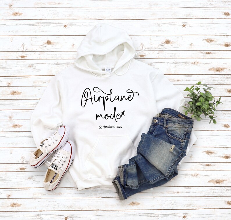 May include: White hoodie with the text "Airplane mode" and a small airplane icon. Below the text is "Maldives 2020". The hoodie is laid out on a white wood surface with a pair of white sneakers and blue jeans.