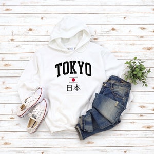 May include: White hooded sweatshirt with black text that says "TOKYO" and a small Japanese flag with the text "日本" underneath. The sweatshirt is laid out on a white wood background with a pair of white sneakers and blue jeans.