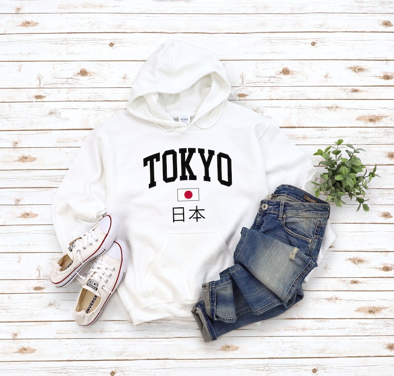 May include: White hooded sweatshirt with black text that says "TOKYO" and a small Japanese flag with the text "日本" underneath. The sweatshirt is laid out on a white wood background with a pair of white sneakers and blue jeans.