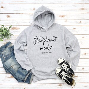 May include: Gray hooded sweatshirt with the text "Airplane mode" and the location "Maldives 2024" printed on the front. The sweatshirt is paired with blue jeans and black sneakers.