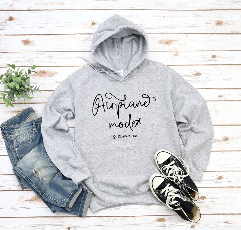 May include: Gray hooded sweatshirt with the text "Airplane mode" and the location "Maldives 2024" printed on the front. The sweatshirt is paired with blue jeans and black sneakers.