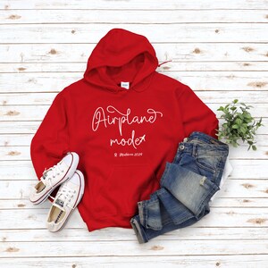May include: Red hooded sweatshirt with the text "Airplane mode" and "Maldives 2029" in white. The sweatshirt is laid out on a white wood background with a pair of white sneakers and blue jeans.