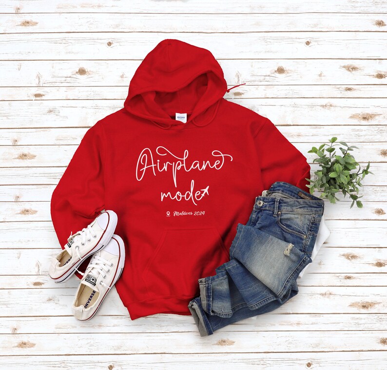 May include: Red hooded sweatshirt with the text "Airplane mode" and "Maldives 2029" in white. The sweatshirt is laid out on a white wood background with a pair of white sneakers and blue jeans.