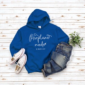May include: A royal blue hoodie with a white graphic that says "Airplane mode" with a small airplane icon and the text "Maldives 2029". The hoodie is laying on a white wood surface with a pair of white sneakers and blue jeans.