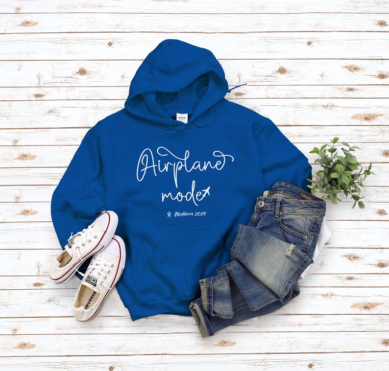 May include: A royal blue hoodie with a white graphic that says "Airplane mode" with a small airplane icon and the text "Maldives 2029". The hoodie is laying on a white wood surface with a pair of white sneakers and blue jeans.