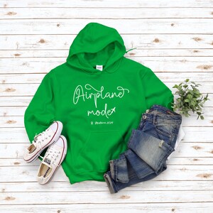 May include: Green hooded sweatshirt with the text "Airplane mode" and the location "Maldives 2023" in white lettering. The sweatshirt is laid flat on a white wood surface with a pair of white sneakers and blue jeans.