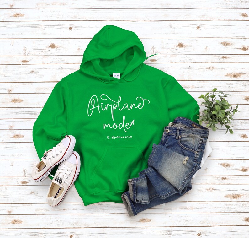 May include: Green hooded sweatshirt with the text "Airplane mode" and the location "Maldives 2023" in white lettering. The sweatshirt is laid flat on a white wood surface with a pair of white sneakers and blue jeans.